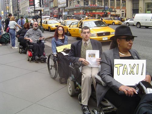 DIA members and other activists are lined up in their wheelchairs on a busy NYC sidewalk holding signs. Behind them are inaccessible yellow taxis at a public taxi stand.