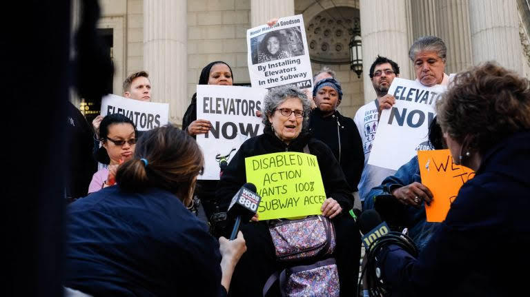 DIA members and other activists protesting for NYC subway accessibility in front of a courthouse, with news reporters holding microphones towards them. One woman is holding a sign that reads "Disabled In Action wants 100% subway access." Behind her are people holding signs that say, "Elevators NOW."